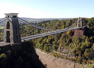 Suspension bridge between two brick-built towers, over a wooded gorge, showing mud and water at the bottom. In the distance are hills.