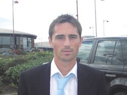 A head and shoulders of a man, wearing a black blazer, white shirt and blue tie.