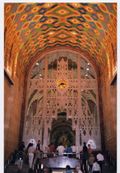 alt=Interior photograph of high, arched ceilings, elaborately patterned with brightly colored designs, while an intricate metal screen partially hides a large mural behind.