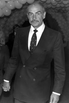 A black-and-white photo of actor Sean Connery, the setting consists of many balloons and two persons are located in the back.