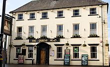 Front of a three-storey building, six windows across, with a large-framed wood door at ground level and a painted sign with the words "THE QUEENS HEAD"