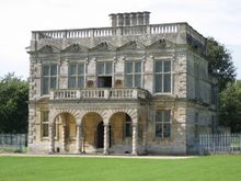 A five-bay, flat-roofed building with two storeys and an arched single-storey portico.  Surrounding the top of the building is a balustrade and chimneys rise from the rear