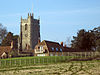 Square stone tower of church with residential buildings. In the foreground grass field contained by wooden fences.