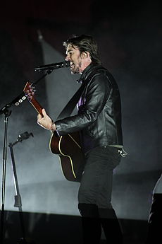 A man in a black shirt, singing into a microphone on a stand and holding a guitar.