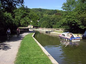 Water in the canal passing over a bridge with stone walls, surrounded by trees. On the left and right are tow paths with cyclists and pedestrians.