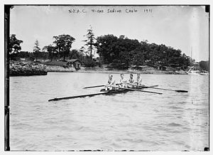 Four men sitting in a long canoe, in front of a wooded camp