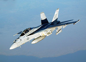 Top view of gray jet fighter banking away from camera. Under the fuselage is an external fuel tank