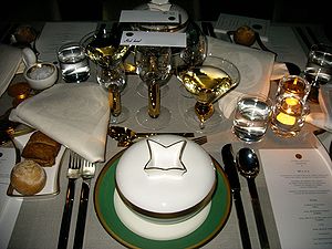 A set table with a white table cloth. There are many plates and glasses plus a menu visible on the table.