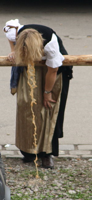 A photograph of a woman with short blonde hair wearing a black dress and standing outside while leaning over a wooden railing and vomiting yellow liquid