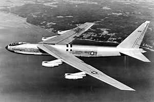 Side, black and white view of an early B-52, which features the bubble canopy. The aircraft is heading to sea, leaving behind the coastline on its right wing.