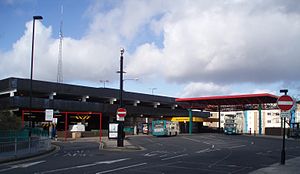 A 1980s multi-storey car park with a large tarmac bus area and light railway station. Two buses can be seen in the station. A red coloured roof is over a section of the bus and metro station and the metro station has its own roof coloured in cadmium yellow.