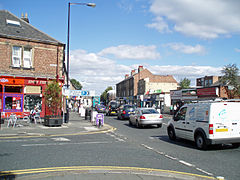 The junction of High Street and Church Road. A terrace of stone fronted buildings, with a curved corner building fronted in white, from the mid-late 19th century. The modern shop fronts are not in keeping the original style of the buildings. It is set back from the road by modern paving. The far section of the building is the old fire station building (formerly Urban District offices).