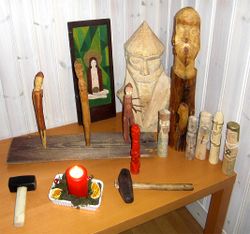 A Heathen altar for household worship in Gothenburg, Sweden. The painted tablet on the back depicts Sunna, the two larger wooden idols Odin (left) and Frey (right), in front of them there are the three Norns, and in the front row a red Thor and other idols. In front of the cult images are two ritual hammers.