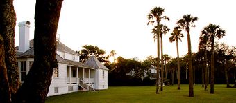 Photo of a sunset over a lawn featuring a large white house to the left, several palm trees on the right, and a wooded area in the background