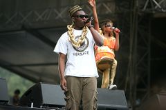 A man wearing an African-style hat, sunglasses, and a large gold chain, and holding a microphone. In the background a woman in brightly coloured clothing and sunglasses is also holding a microphone and is waving one hand in the air