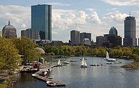 Skyline of a city on a river. Two buildings are much taller and more prominent than the rest; one of the large buildings is a skyscraper with a blue-tinted, all-glass facade, and the second is a large rectangular tower with a steel latticework facade and a tall antenna mast on its roof.