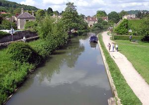 Wide stretch of still water with a narrow boat on it. Tow path on the right and trees and houses on the left.