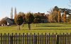 Stone buildings seen behind trees and grass area with wooden fence in the foreground.