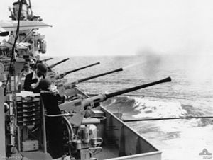 The flank of a ship. Several long-barrelled guns are aimed over the side, and are being operated by sailors. One of the guns has just fired, with a cloud of smoke issuing from the barrel.