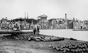 A single soldier stands among cannons and cannonballs across a river from the ruins of a city.