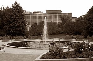 a large, flat-roofed building, partially obscured by trees, with a fountain in a circular pond in the foreground