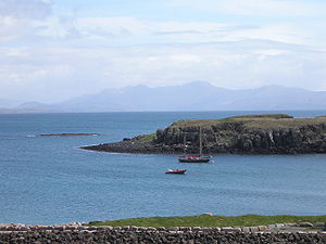 A two-masted yacht and a RIB lie at anchor on a sunny day off  a grassy shore, with low cliffs beyond. A skerry lies further offshore to the left with high hills in the distance.