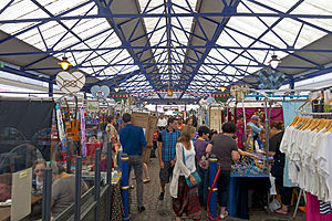 An interior of a building with a translucent glass roof supported by blue-painted steel latticework. On the main floor are a number of different stalls with customers inspecting various wares.