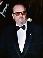 A Caucasian man is seen wearing sunglasses, a black suit over a white collared shirt and a black bowtie.