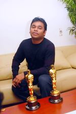 A color photograph of a man sitting on a sofa, with two golden statuettes on the table in front of him.