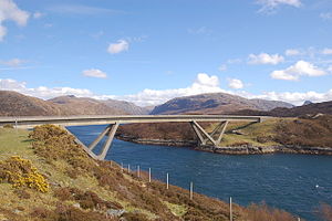 A concrete road bridge spans a body of deep blue water with brown hills beyond