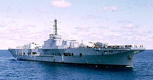 A small aircraft carrier underway in calm, open seas. Structures have been built on the ship's flight deck, and additional cranes have been installed. No aircraft can be seen, and a large number of people are clustered around the forward end of the flight deck.