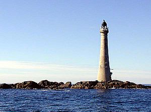 A tall lighthouse stands on low rocks under a blue sky.