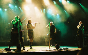 Four singers perform in the spotlights on stage. One is a female in shorts, a gauzy beige skirt and a leather belt, and plays the fiddle,  The males are wearing black sleeveless shirts, black pants and have military-looking gear. A rock band is in the background.