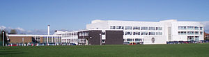 A large group of school buildings. The white building in the background was built in the early 21st century whereas the darker buildings on the left were built in the 1960s or 1970s. In the foreground the front grass field. Since the photo was taken the grass has been replaced by an artificial turf pitch.