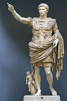 A marble statue of the Emperor Augustus. He stands with one arm raised as if in command. Augustus is depicted as a man of about thirty five, with short hair and clean shaven. He wears Roman military uniform of a breast plate, leather accoutrements and a cloak over a short tunic. The breastplate is decorated with symbolic figures. As a work of art, the statue displays high technical mastery.