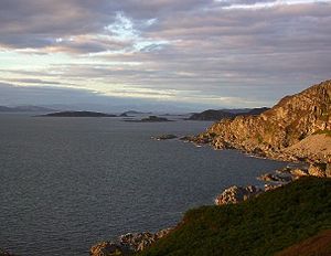  A sunlit rocky promontory at right and a calm, dark sea at left. Small dark islands pepper the waters in the distance