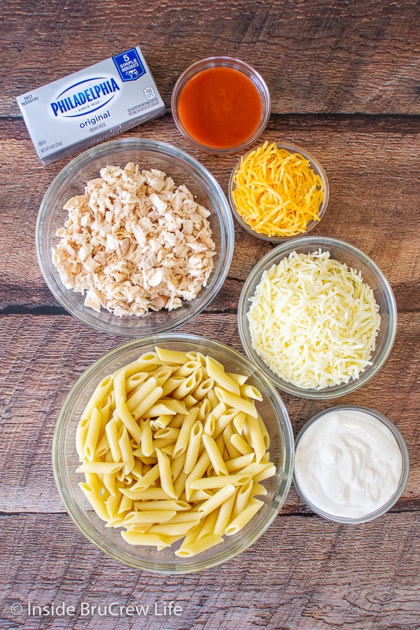 Bowls of ingredients to make buffalo chicken pasta bake on a wooden board.