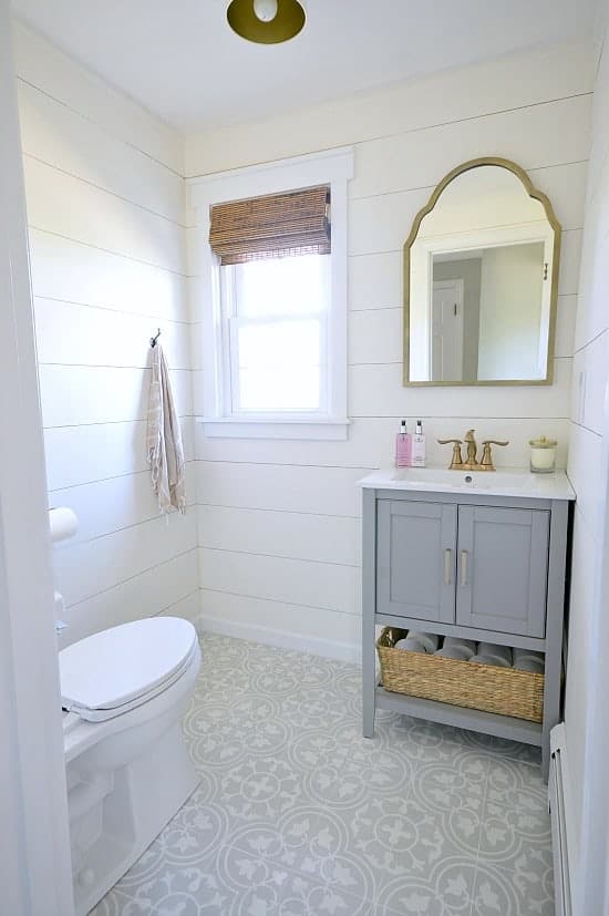 A bathroom with grey vanity, gold mirror, shiplap walls and a small window with rolled-up wooden screen