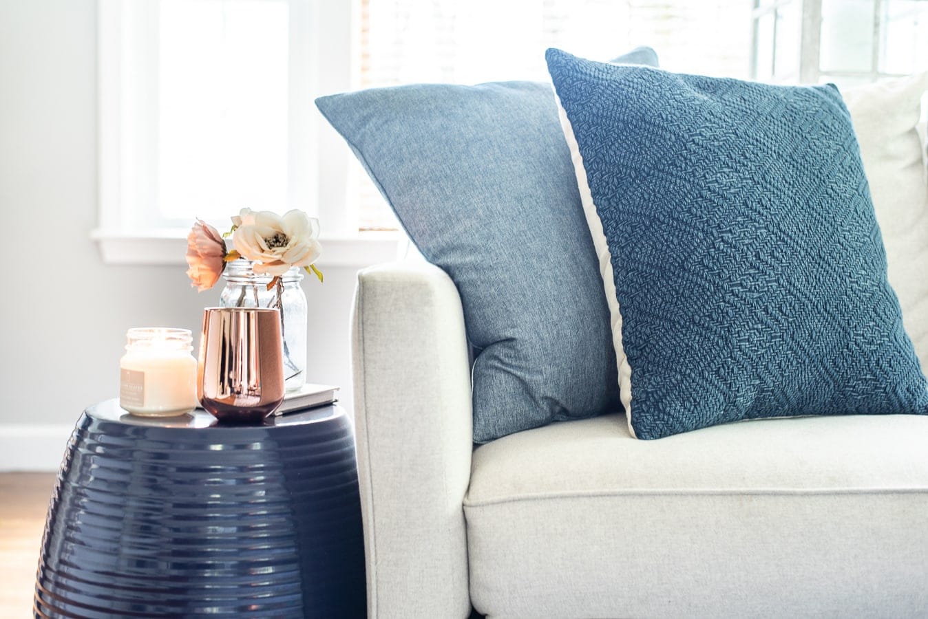 The corner of a light grey couch, with two dark blue pillows, near a metallic blue side table and a copper vase of flowers