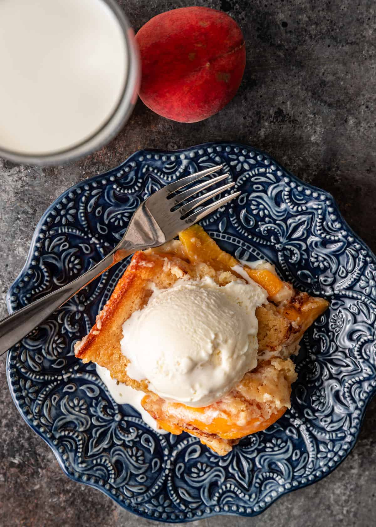 overhead: a slice of peach cobbler with vanilla ice cream on top on a blue plate with a fork