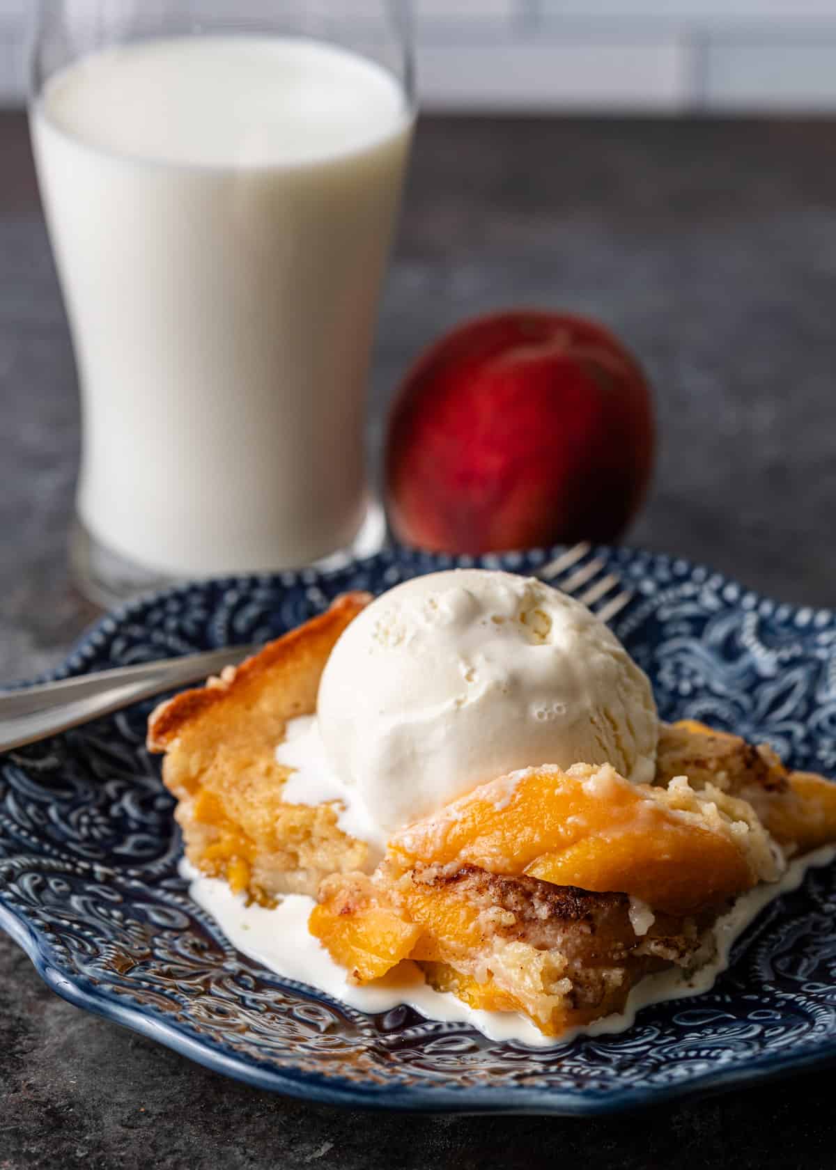 closeup: a slice of my peach cobbler recipe with vanilla ice cream on top with a glass of milk and a peach in the background