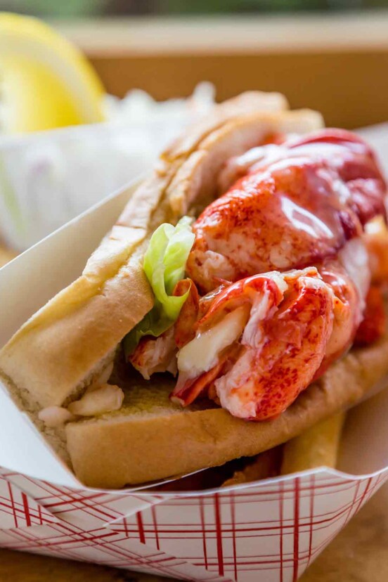 A lobster roll in a cardboard sleeve with a little lettuce and a lemon wedge in the background.