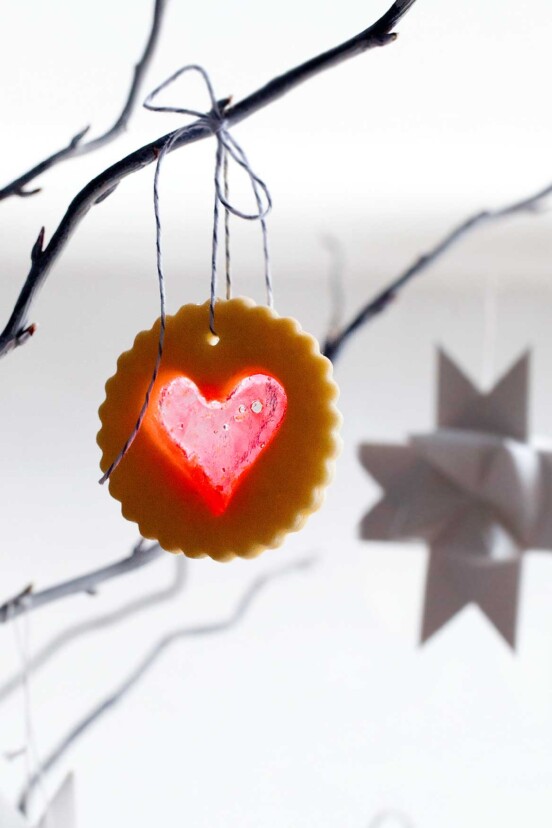 A scalloped round sugar cookie with a heart-shaped windowpane cutout hanging from a tree branch.