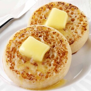 Two English crumpet on a white plate, each topped with a pat of butter.