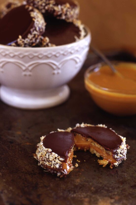 A white bowl filled with alfajores, and one halved one in front of the bowl with a bowl of dulce de leche beside