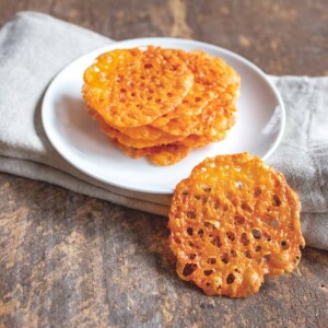 A white plate sitting on a folded napkin on a wooden table, with a pile of cheese crisps on top and one leaning against the plate.