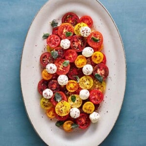 A large white platter filled with halved various colored cherry tomatoes, labneh balls, olive oil, and a sprinkle of spices.