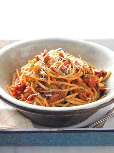 A brown and white pottery bowl filled with bucatini all'Amatriciana, topped with pecorino and parsley.