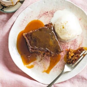 Sticky toffee pudding with vanilla ice cream on a white plate with a fork, beside a small bowl of extra toffee sauce.