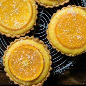 Five small tarts filled with orange curd and topped with candied orange slices on a cooling rack.